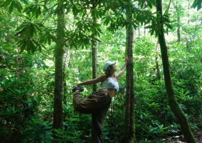 Nazan is in a supported warrior 3 pose in a forest, surrounded by trees. She has a cap on, and is smiling.She is using the tree for support. The background is lit with summer sunlight and tall trees.