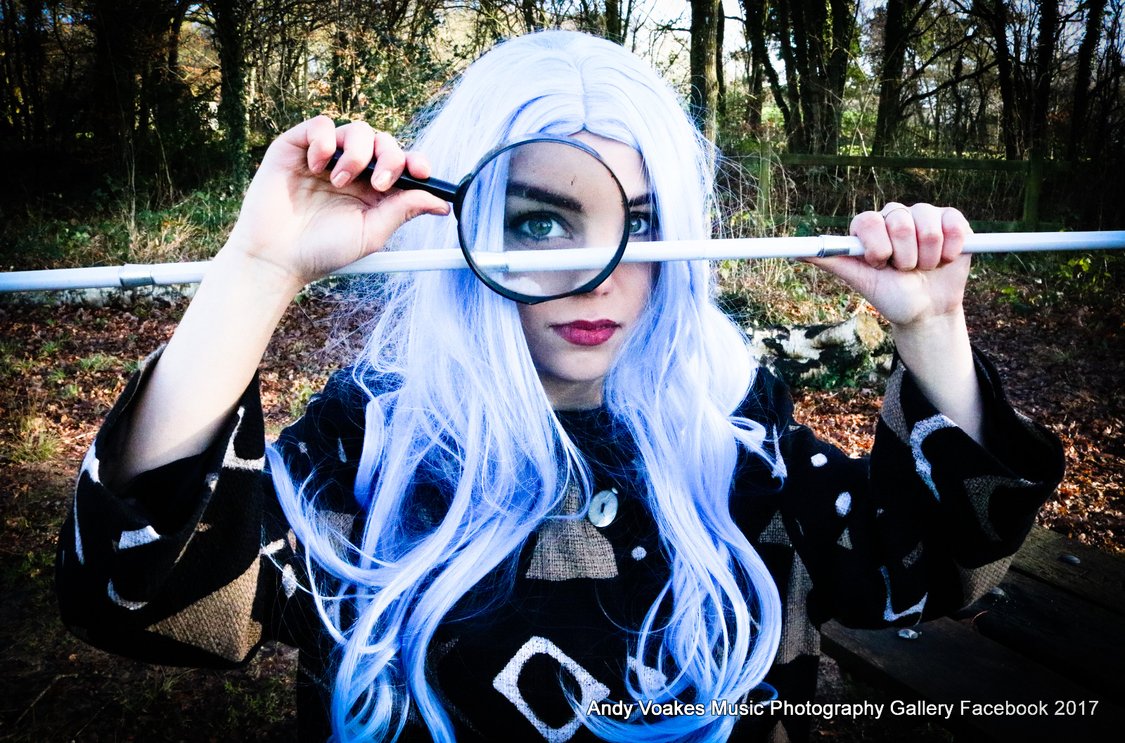 Image shows Nazan, with a long wig on, a white cane being held by herself across her face, and a magnifying glass in front of one eye. The photo is sharp, bright and black and white. It was taken by Andy at Big Bear Photography.