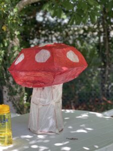 This image shows a huge paper mache mushroom! (Crafts section). It has a white stalk, and red head with white polka dots -- it is set on a plastoc white table with a backdrop of green leaves (the background is blurred and the mushroom is the focal point, pretty much in the center of the image).