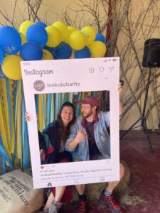 This image shows Will & Naz with a fake photo cut out where it looks like a post on instagram. They are holding the template up and smiling behind it. They have a backdrop behind them, with balloons! 