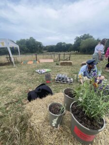 This image shows pot planting with Will! In the forefront, there are a couple of plants on a hay bale, and behind Will is talking to a child (out of view), showing her how how to. pot a plan, and soil stuff etc. Will is smiling, with a fakr RAF hat on! There are hay bales around him, compost and pots. 