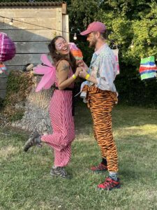 This image shows Nazan and Will are holding hands in costume. Will has his face painted as a tiger, and is half wearing a tiger onsie. Nazan is smiling, with a butterfly face paint on her face, and has wings! 