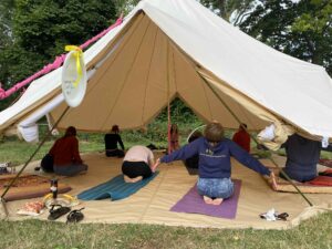 This image shows Nazan teaching yoga to a group of staff and parents/carers. This is set in a large yurt. The backdrop displays trees, and the yurt is installed onto grass. People are practicing in the yurt in various yoga poses on mats.
