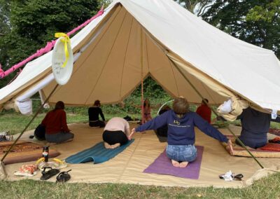 The image shows Nazan teaching yoga to a group of staff and parents/carers. This is set in a large yurt. The backdrop displays trees, and the yurt is installed onto grass. People are practicing in the yurt in various yoga poses on mats.