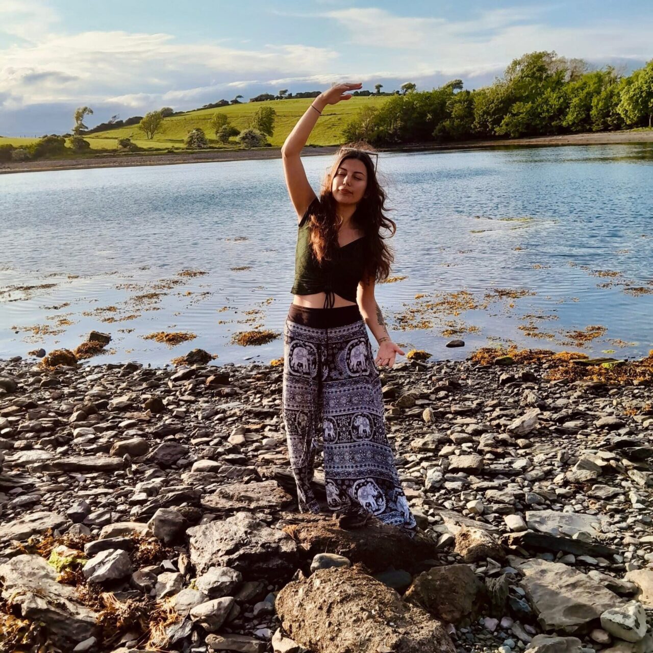 This image shows Nazan in a dancing posture, standing still with one arm up in the air and one arm off to the side. She is placed on top of big rocks next to a shore. Her hair is blowing in the wind. In the distance there are trees, and hills and it looks like a sunny day.