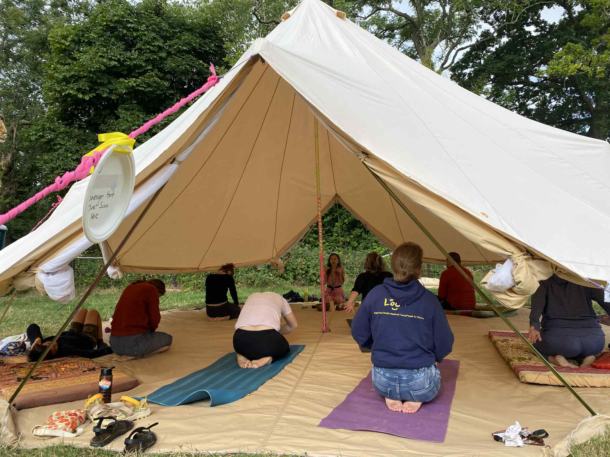 This image shows Nazan teaching a group of people at LOOKFEST. LOOKFEST is a festival for blind children, teenagers, and tehir families. The class is set in a large yurt, filled with people. (Maybe around 8-10 people). The people are on mats, practicing various poses, as Nazan is placed central, teaching from the back of the yurt (large tent). 