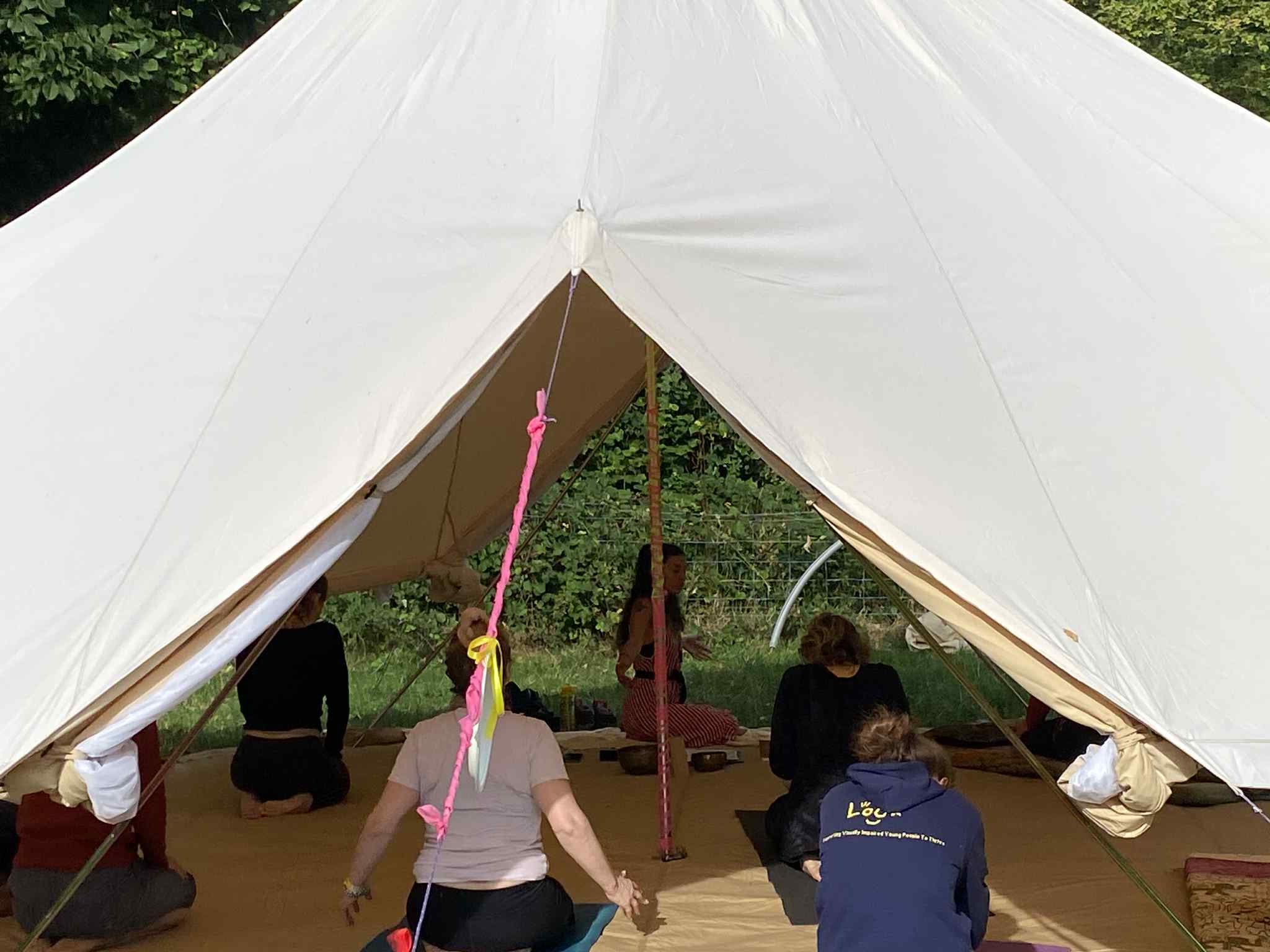 This image shows Nazan teaching a group of people at LOOKFEST. LOOKFEST is a festival for blind children, teenagers, and tehir families. The class is set in a large yurt, filled with people. (Maybe around 8-10 people). The people are on mats, practicing various poses, as Nazan is placed central, teaching from the back of the yurt (large tent). This is a close up photo, as if peaking through the small opening of the yurt.